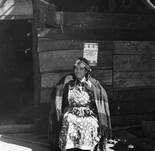 Temuco: Mujer mapuche (Amiga de Cardoso) sentada afuera de su ruca que tiene en la pared un afiche que dice: &quot;Piensen compañeros que el Consejo Comunal es el único camino&quot;. Sentada afuera de su ruca tocando un kultrú, con sus collares de plata; desta