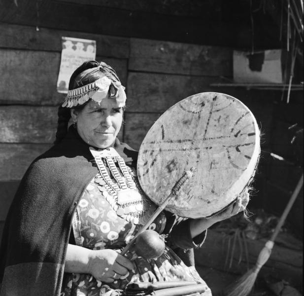 Temuco: Mujer mapuche (Amiga de Cardoso) sentada afuera de su ruca que tiene en la pared un afiche que dice: &quot;Piensen compañeros que el Consejo Comunal es el único camino&quot;. Sentada afuera de su ruca tocando un kultrú, con sus collares de plata; desta